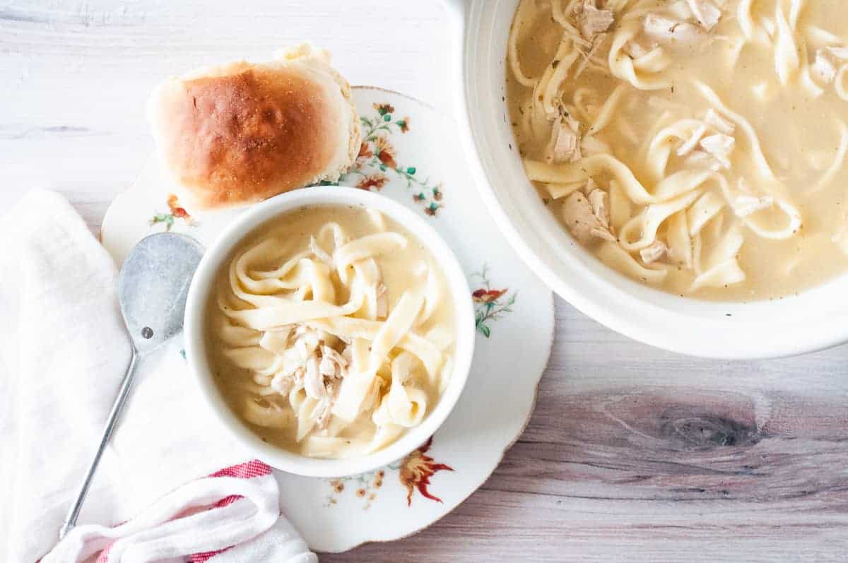 A bowl of chicken and noodles next to a tureen of soup.