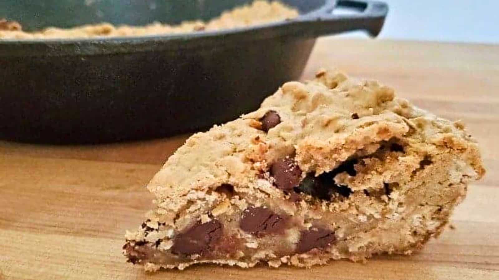 Image shows A piece of chocolate chip skillet cookie in front of a cast iron pan.