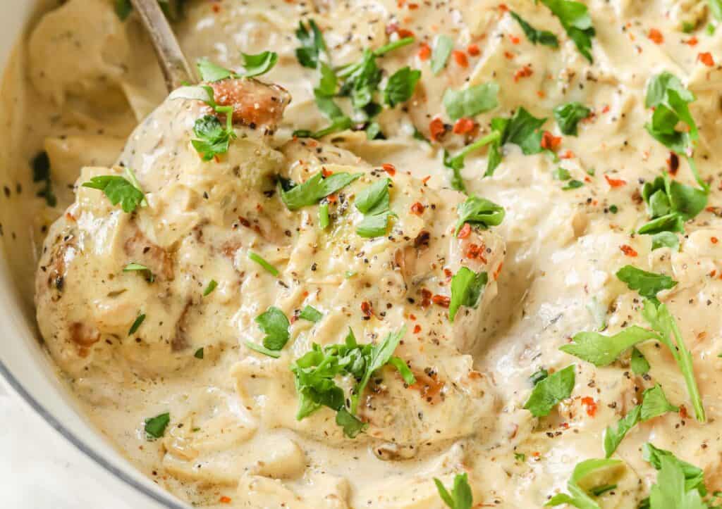 Chicken thighs in cream sauce with artichoke hearts in a pan with a spoon.