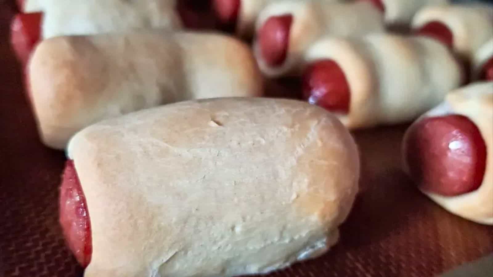 Image shows Hot dogs wrapped in crescent roll dough on a baking sheet.