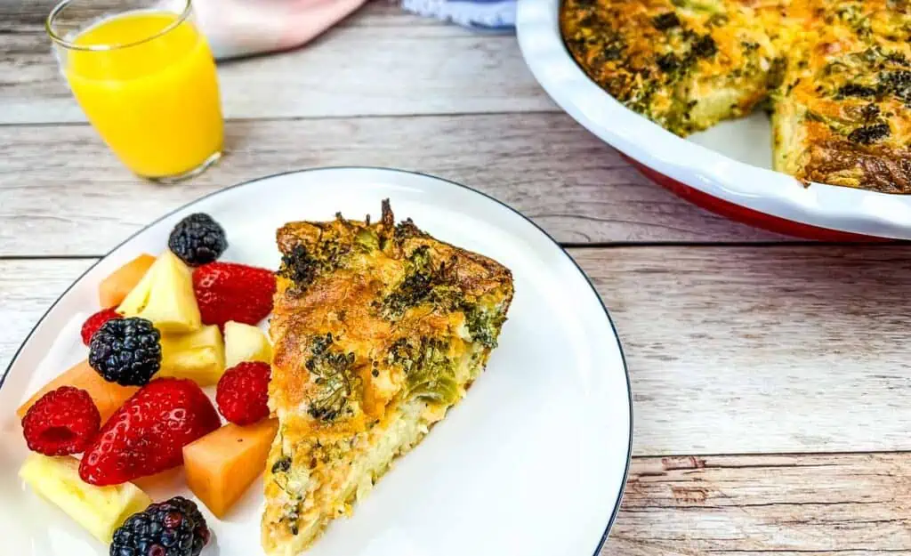 A slice of Broccoli 3-Cheese Impossible Quiche on a plate next to a glass of orange juice.