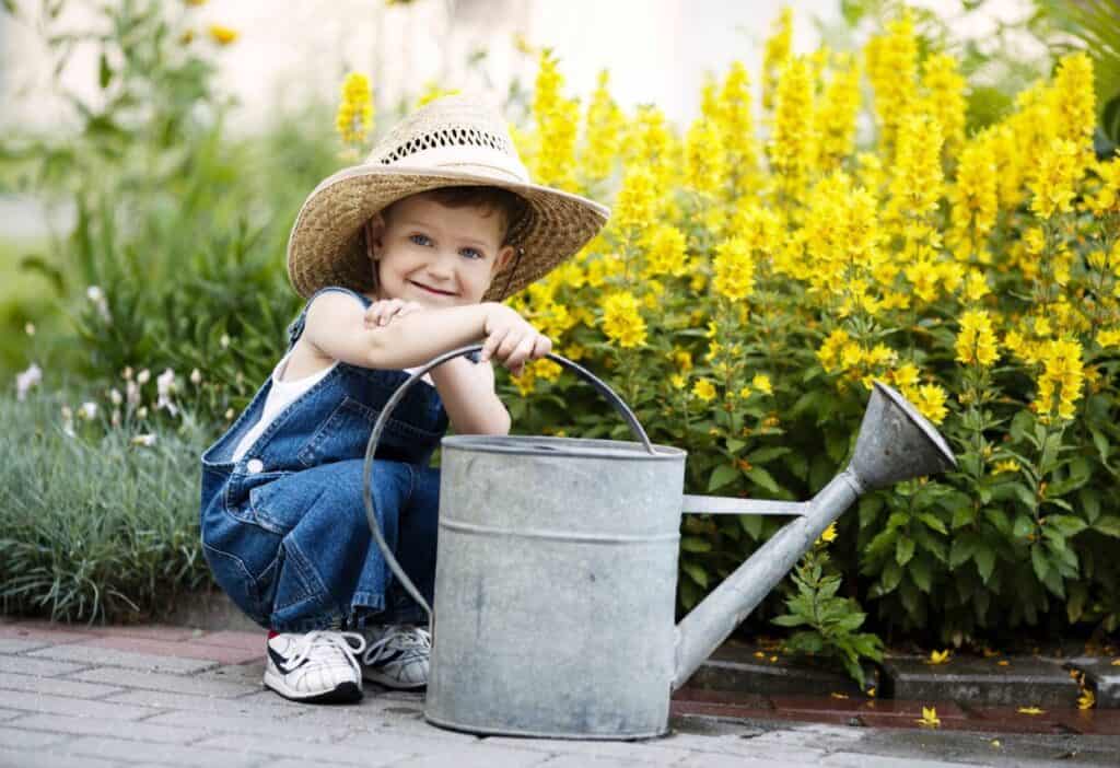 A little boy in overalls sitting next to a watering can.
