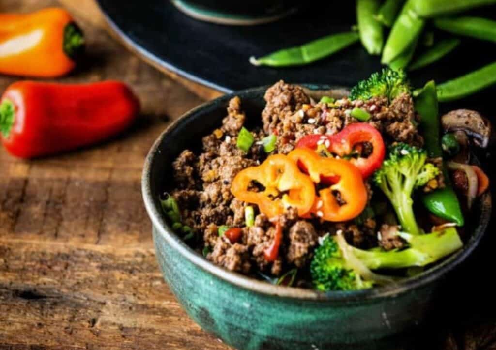 A bowl of beef, broccoli and peppers on a wooden table.
