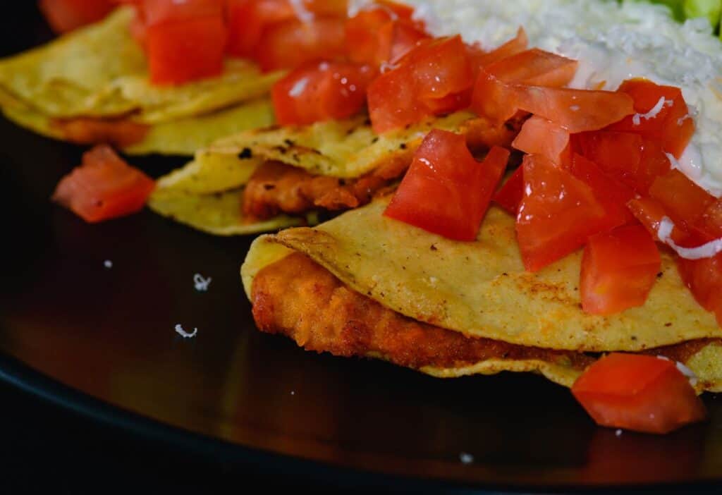 Image shows A black plate with a potato and chorizo tacos topped with tomatoes and crema.