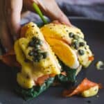 A person holding an eggs benedict with salmon and capers on a plate.