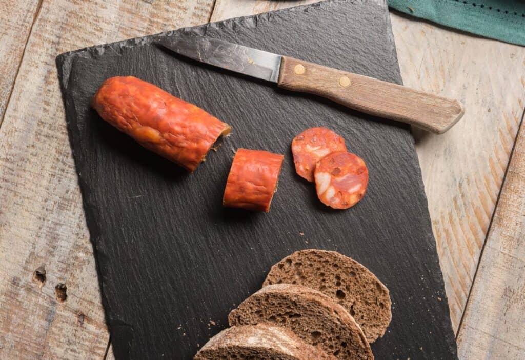 Image shows Spanish chorizo and bread on a slate board with a knife.