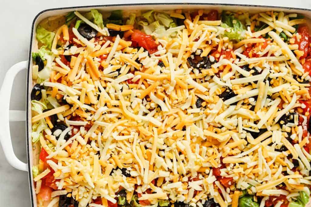 A pan of taco dip with cream cheese, vegetables, and shredded cheese.