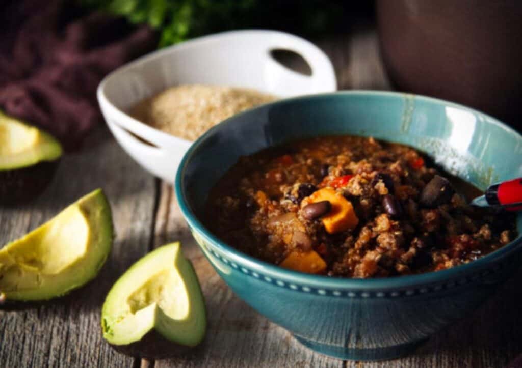 A bowl of chili with beans and avocados on a wooden table.
