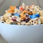 Image shows white chocolate party mix piled into a white bowl on a wooden table.