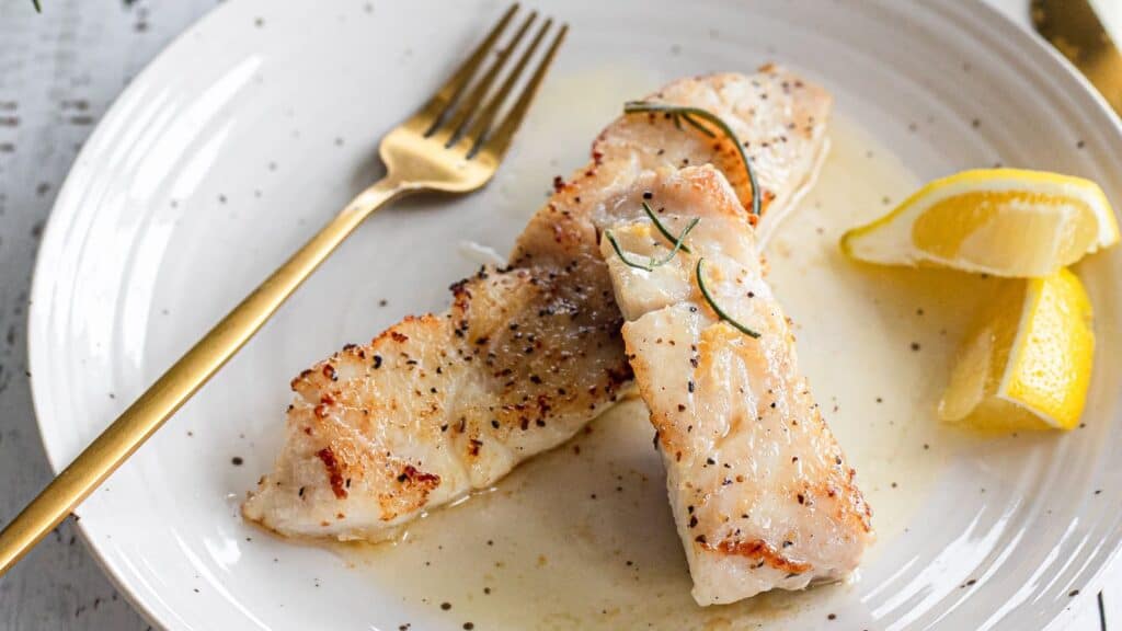 White fish fillets on a white plate with lemon wedges and fork.