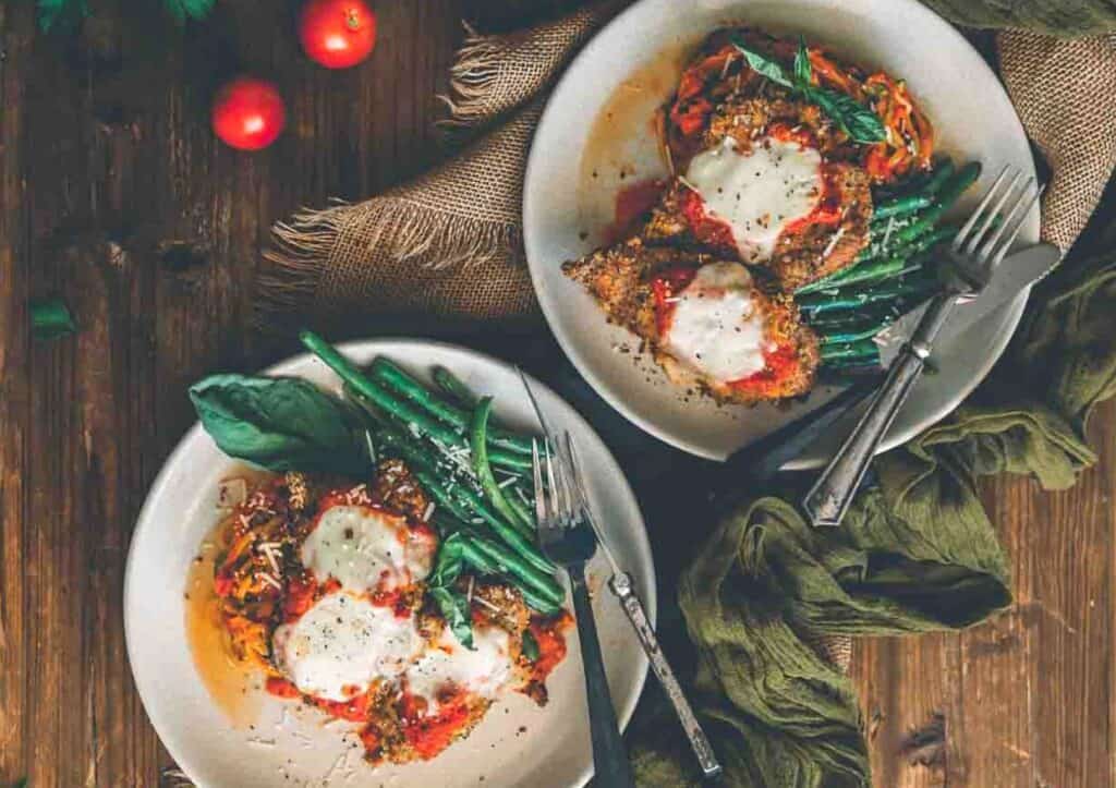 Two plates of veal parmesan and green beans on a wooden table.