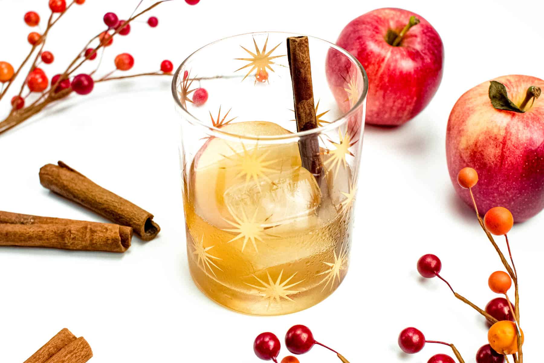 A rocks glass with large square ice cube and golden liquid is garnished with a cinnamon stick and apple slice. Around it on a white background sit apples, cinnamon sticks and sprigs of red berries