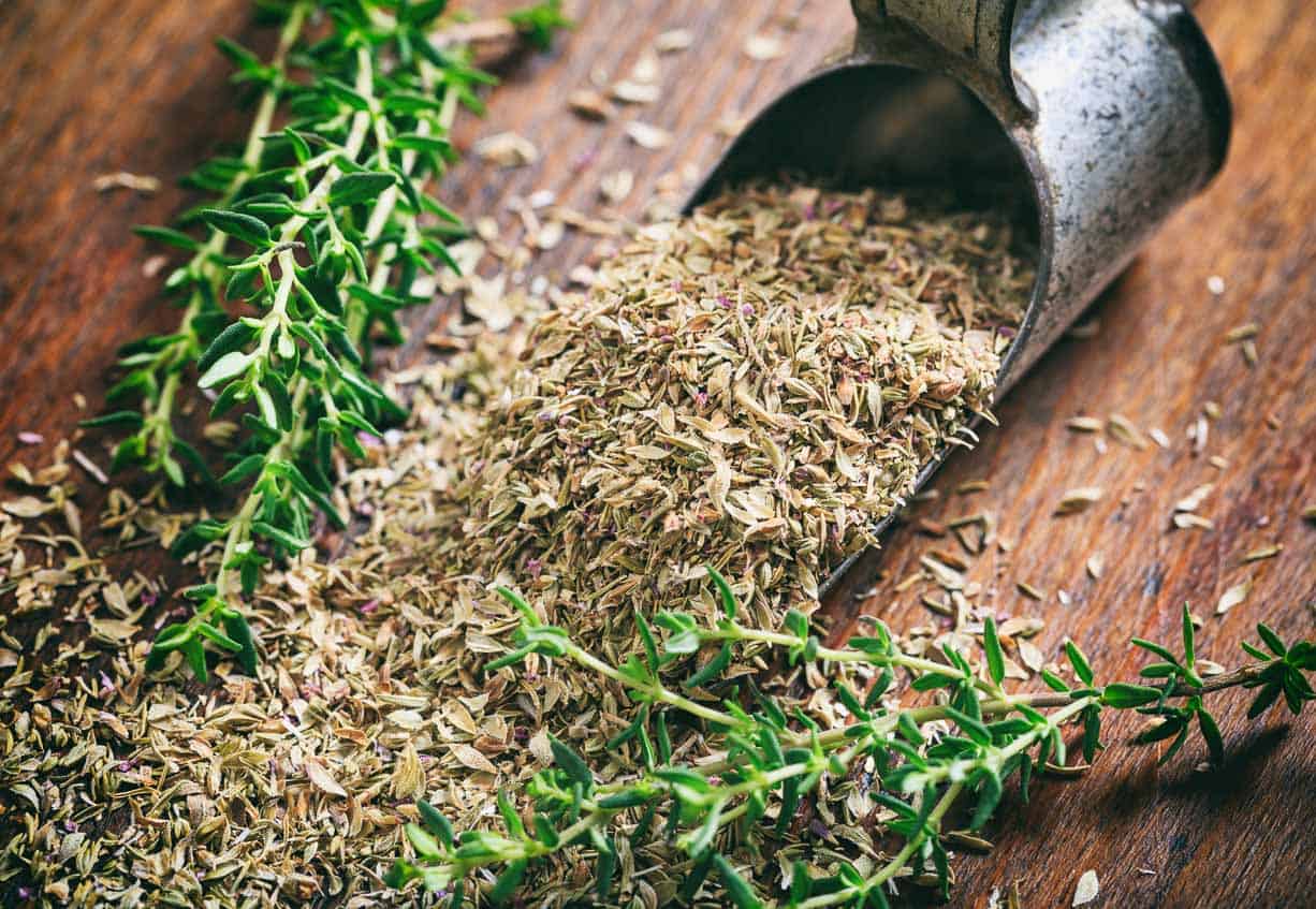 A spoon full of fresh and dried thyme leaves on a wooden table.