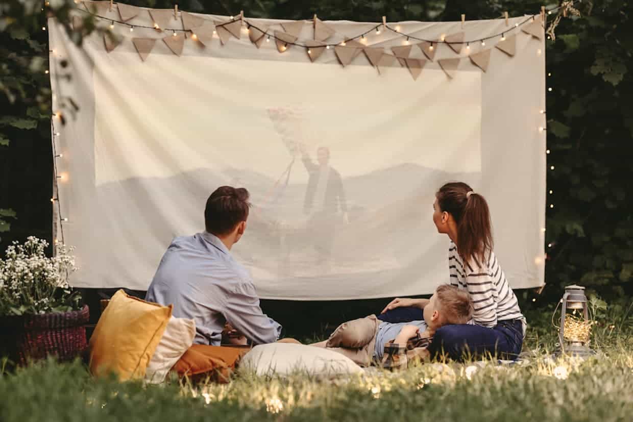 Parents and son sitting on blanket with pillows and watching family camping movies on a big cloth outdoors.