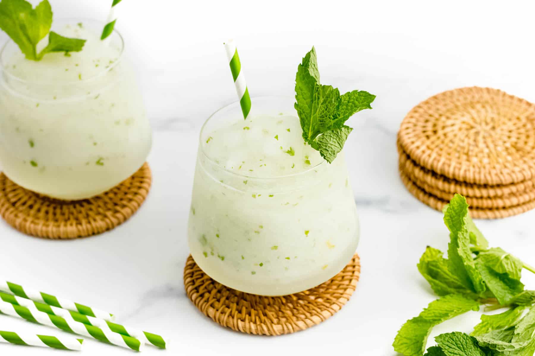 Frozen Mojitos alongside herb sprigs and green and white striped straws.