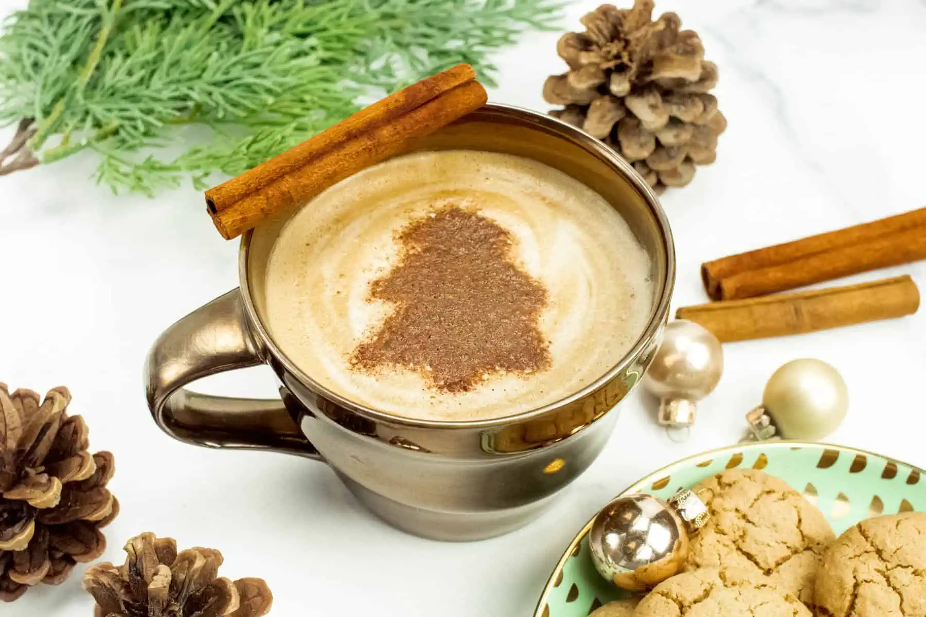 A decorative gold mug with a Christmas tree garnish is surrounded by cinnamon sticks, gingerbread cookies, pinecones and evergreen boughs.