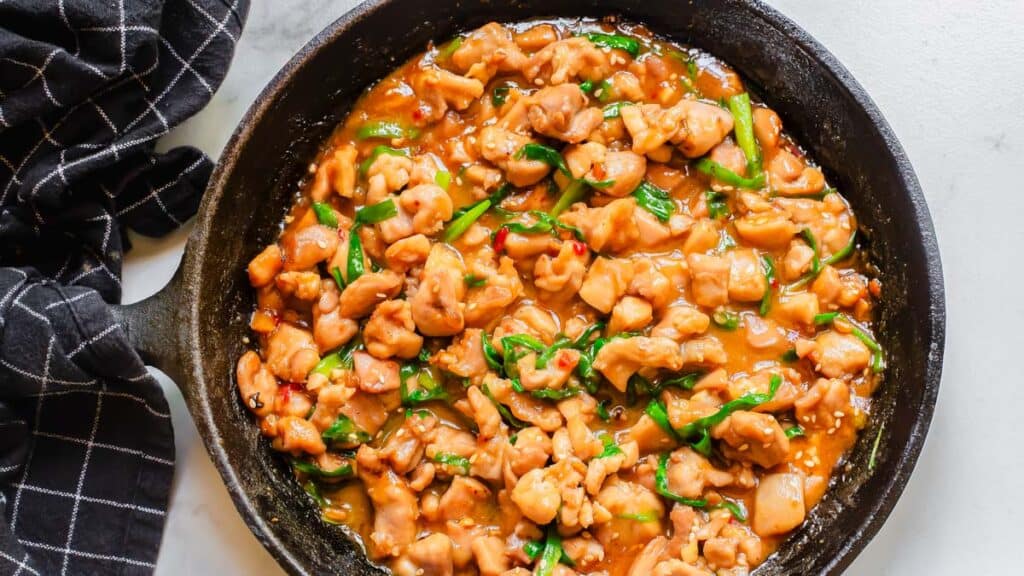 Chinese gochujang chicken in a skillet on a table with black kitchen towel wrapped around the handle.