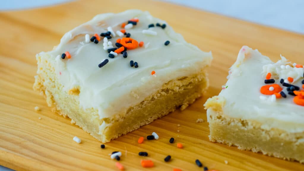 Two sugar cookie bars with sprinkles on a wooden cutting board.