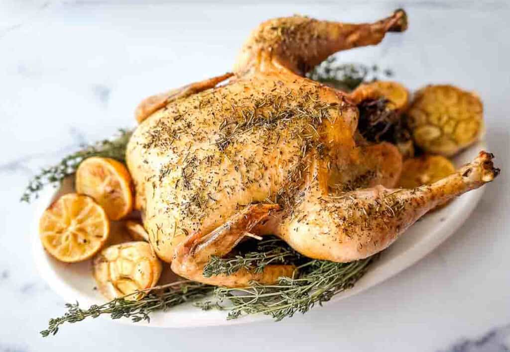 A roasted chicken with thyme and lemons on a white platter.