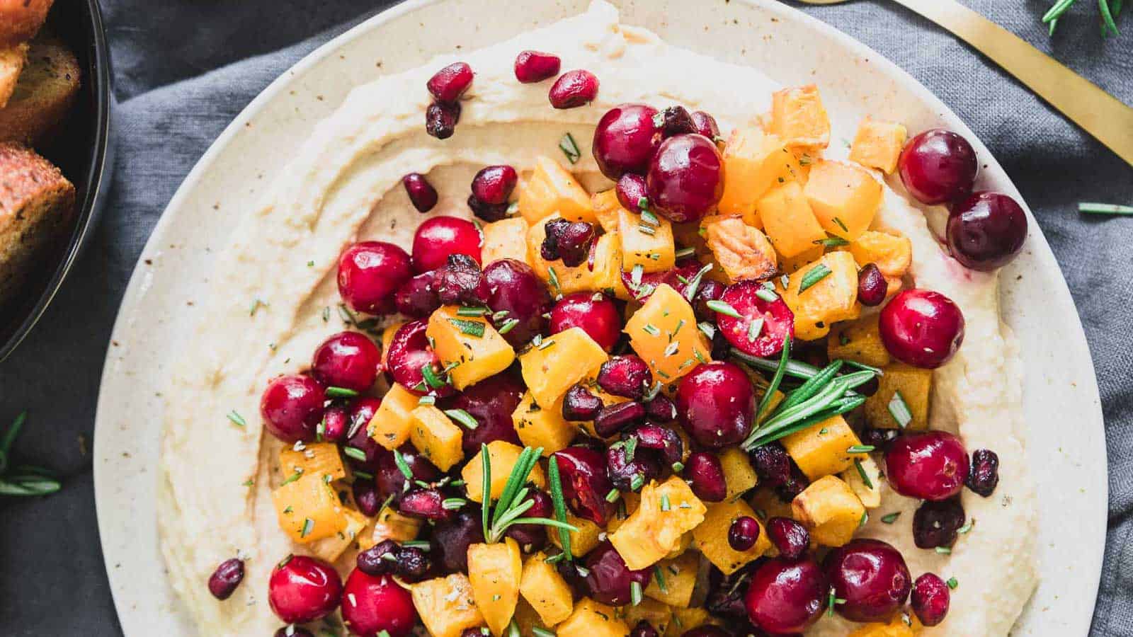 A plate of hummus topped with butternut squash, cranberries and rosemary.