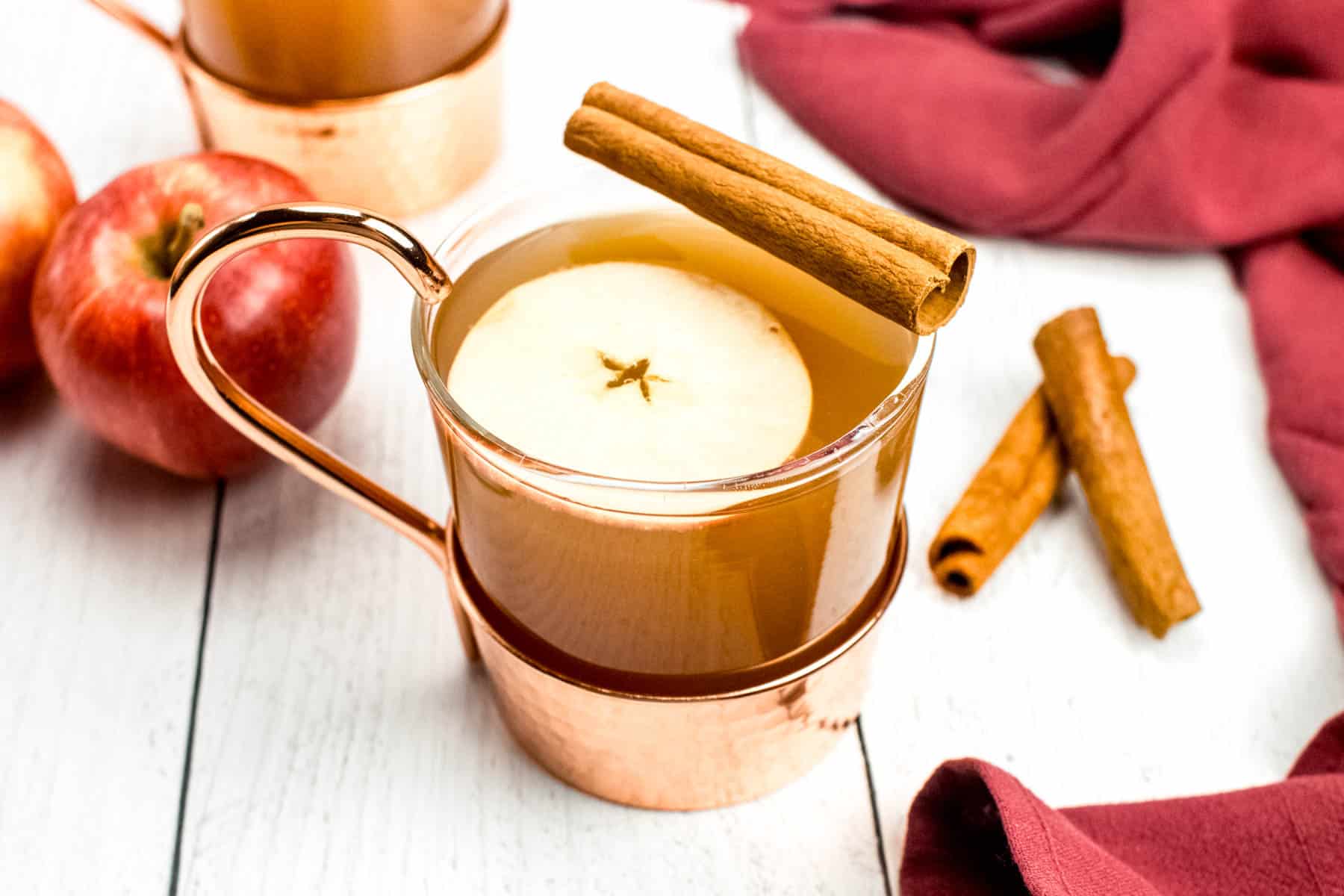 A cup of apple cider with cinnamon sticks and apples.