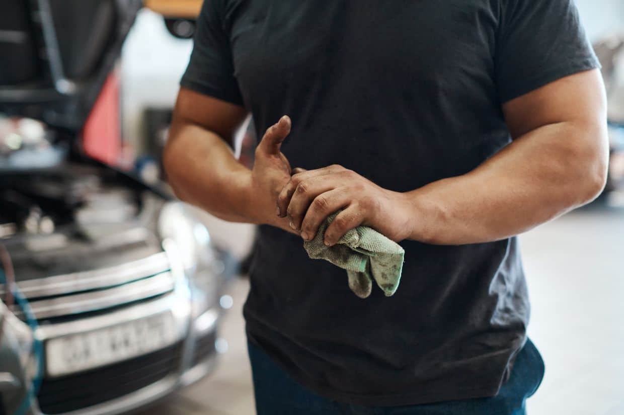 A man in a black shirt is standing next to a car in a garage, seeking help on getting gas smell off hands.