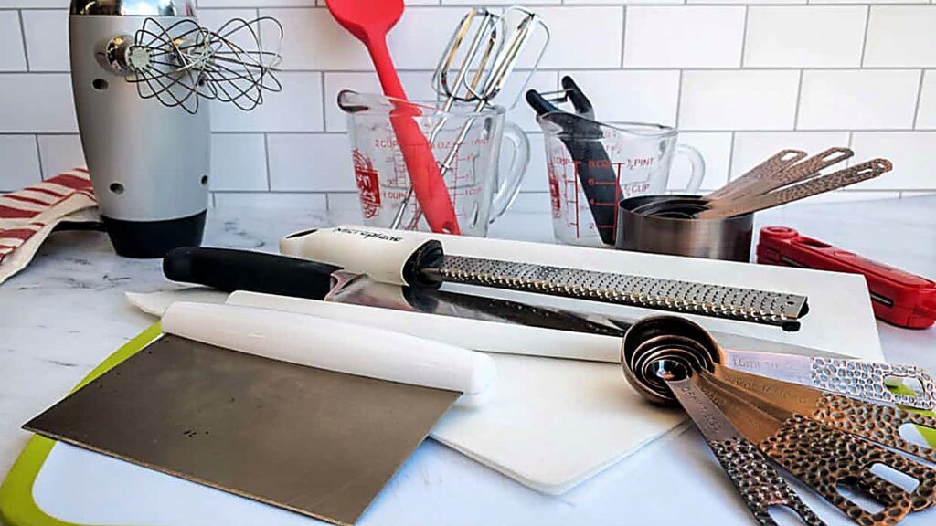 A variety of essential kitchen tools, including measuring cups, spoons, knives, a microplane, spatula, and a hand mixer, are arranged on a countertop with a white subway tile backsplash.