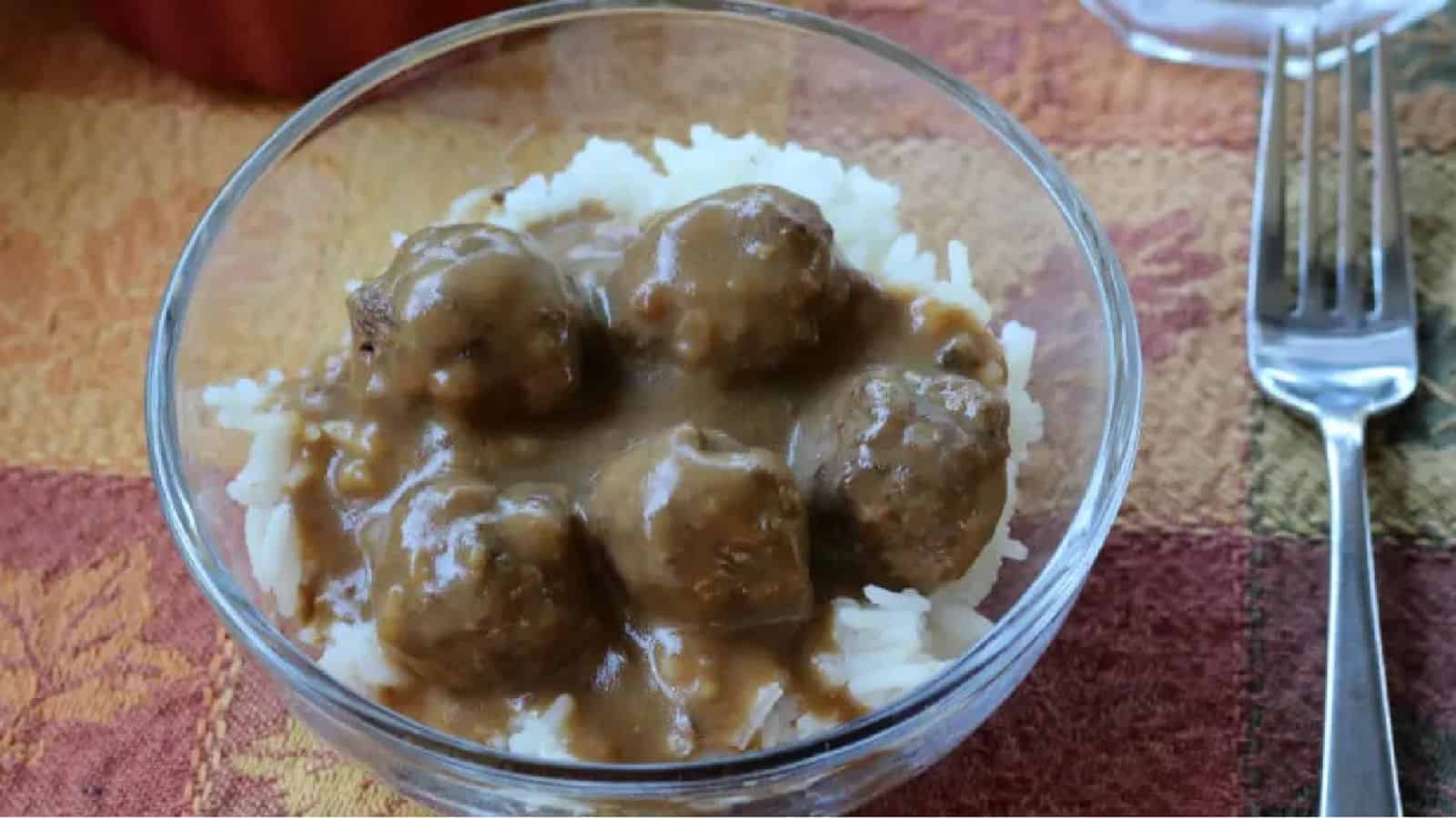 Glass bowl of meatballs and gravy over rice, fork to the side, grapes in the background.