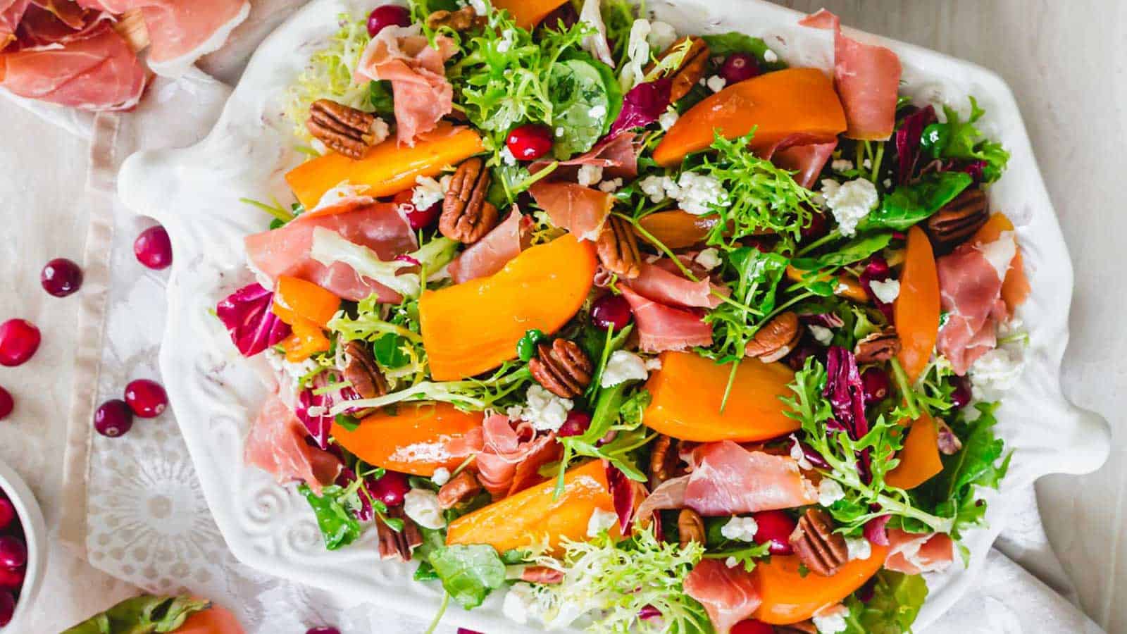 Persimmon salad with greens, prosciutto, cranberries and pecans on a white serving platter.