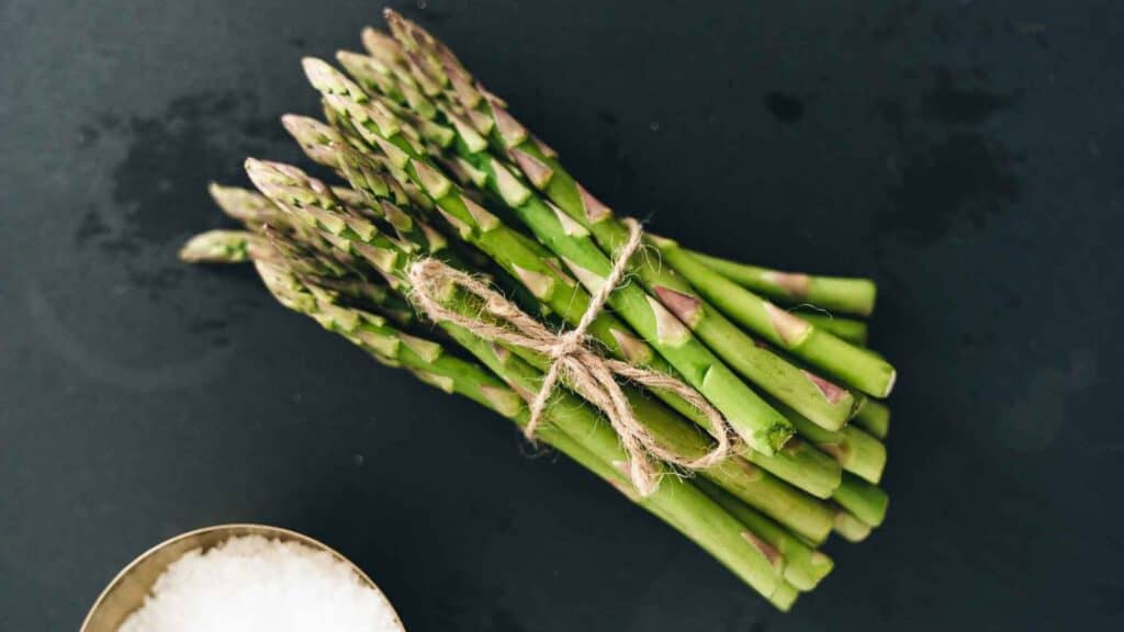 A bundle of fresh green asparagus spears tied with twine, placed on a dark surface next to a small bowl of coarse salt.