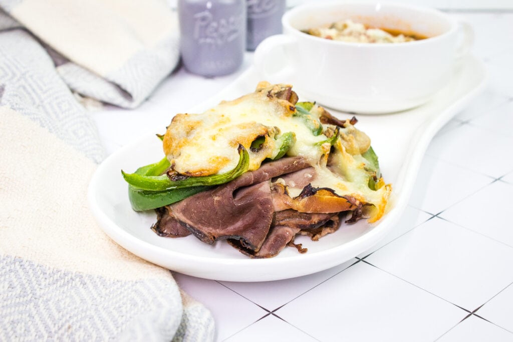 A white plate with a Philly Cheesesteak Stack and a bowl of soup.