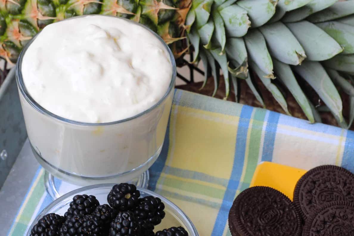 Glass bowl of pineapple dip, blackberries, cookies, and a pineapple in the background.