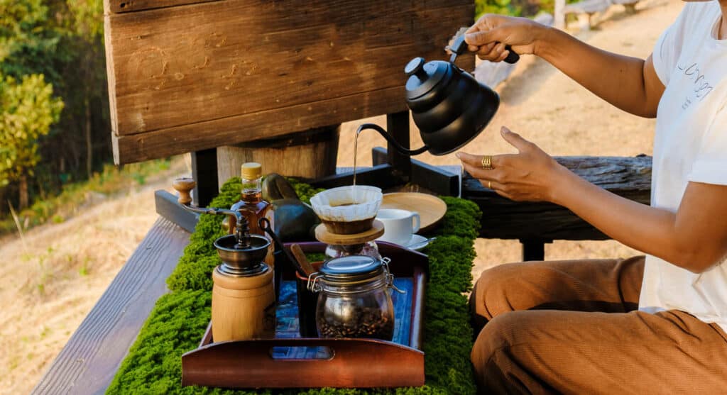 A woman is pouring coffee on a wooden table outside.