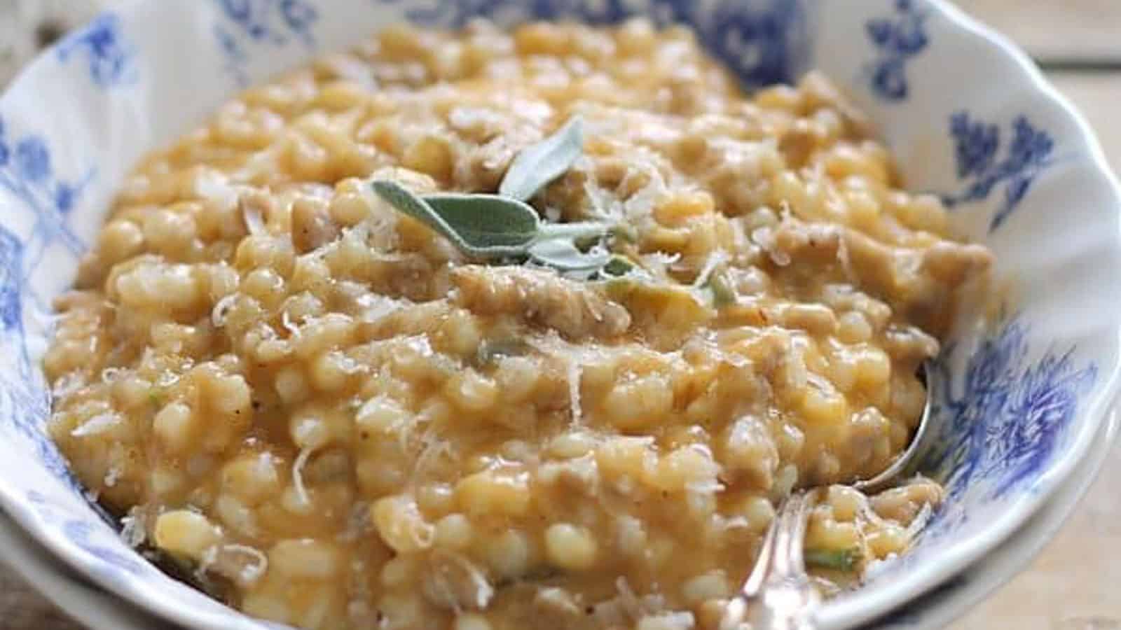 Creamy pumpkin barley with sausage garnished with fresh sage leaves and grated parmesan in a bowl with a spoon.