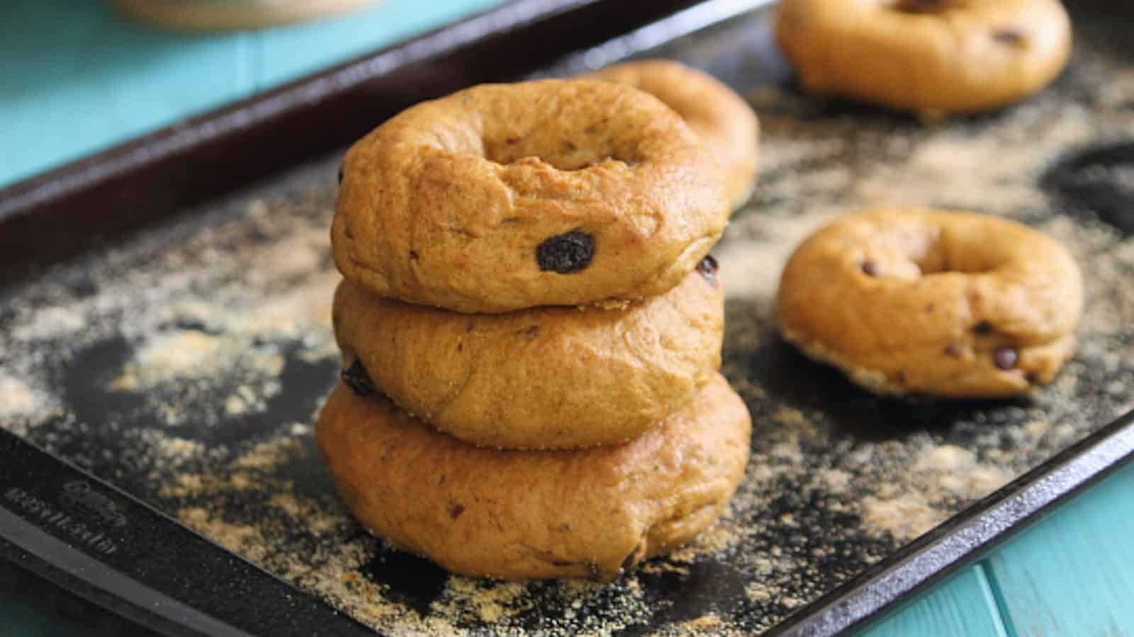 Three pumpkin raisin bagels stacked on a baking sheet dusted with cornmeal.