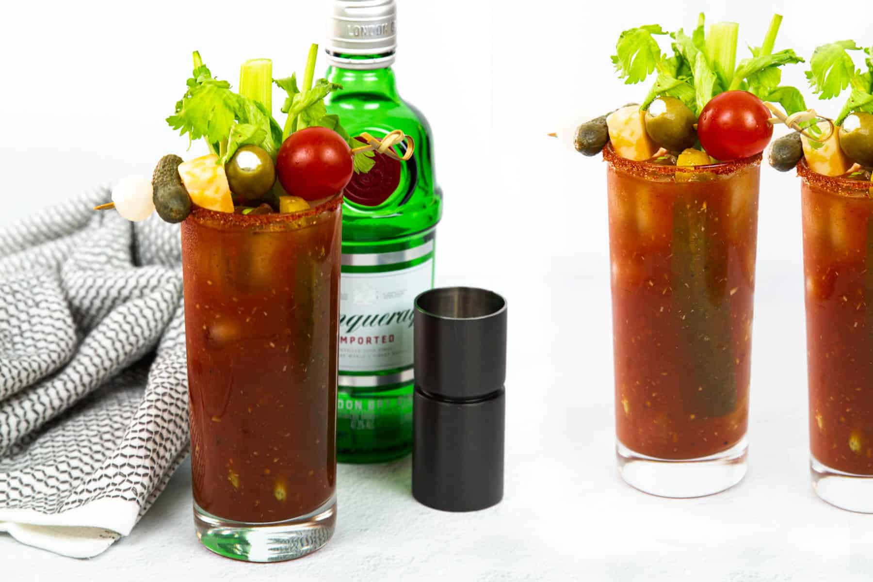 Three glasses of bloody mary with vegetables and a bottle of gin.