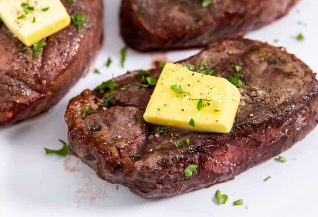 Three steaks with butter and herbs on a white plate.