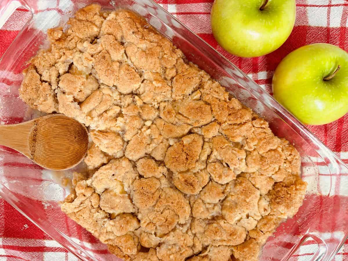 Apple crisp in a glass dish with apples and a wooden spoon.
