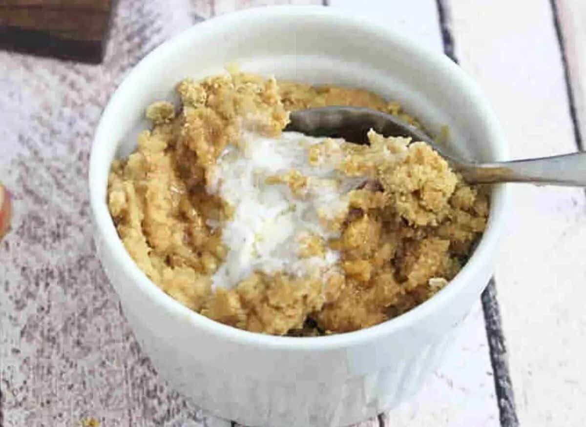 Apple mug cake in a white ramekin with coconut milk on top and spoon in it and apples and cinnamon around it.