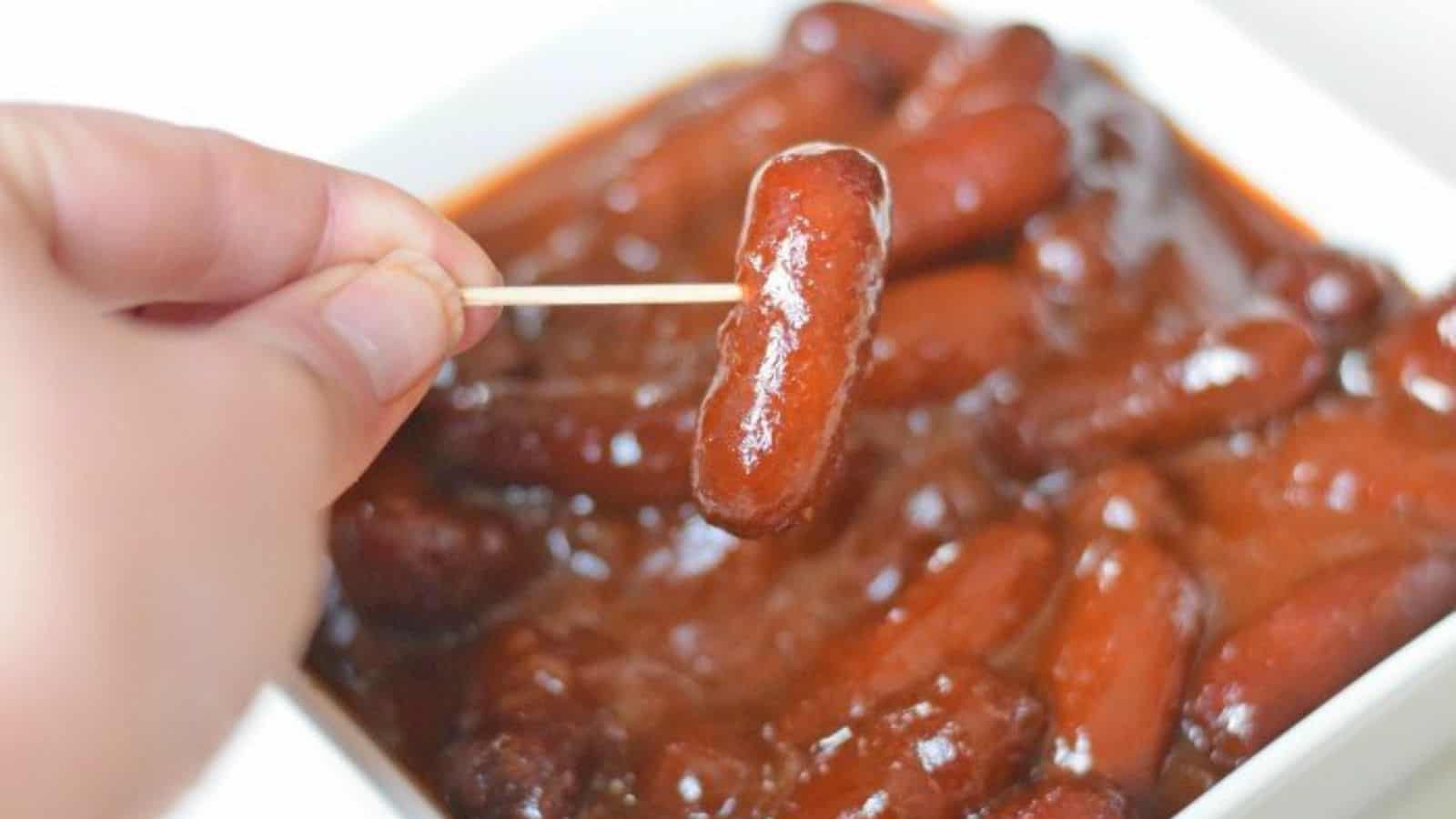 Image shows A person is holding a toothpick with a little smokie over a bowl filled with more bbq bourbon little smokies.