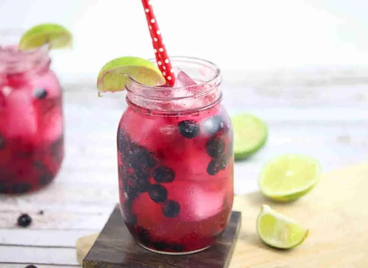 Blueberry mocktail in a mason jar with limes on the side of the jar and red straws with the jar on a wooden coater and cutting board and ingredients around it.