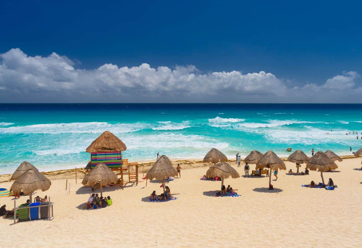 Image shows A beach with thatched huts and umbrellas in front of the ocean.