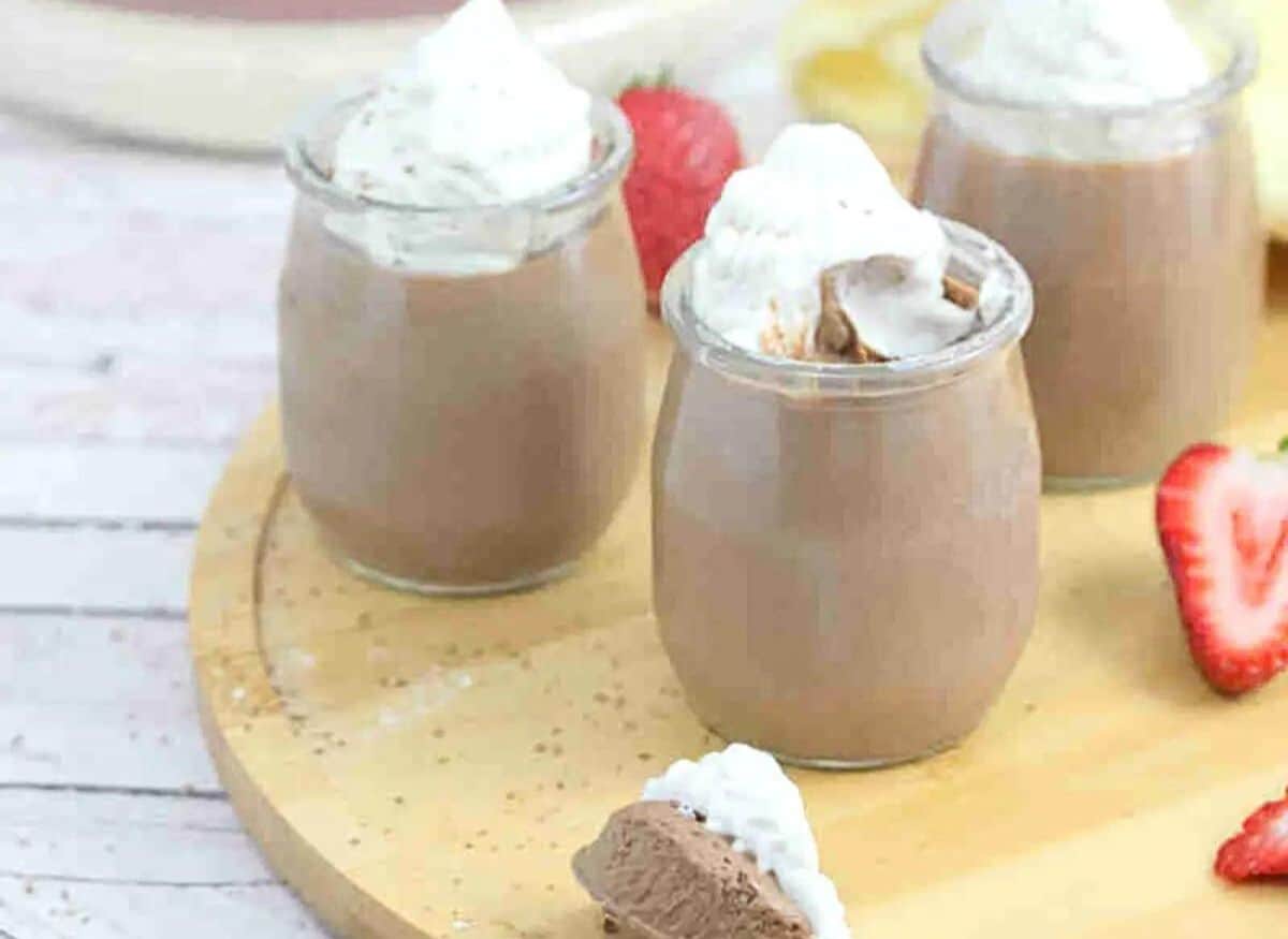 Three glass jars of chocolate protein mousse topped with coconut whipped cream on a wooden cutting board with strawberries around them and a spoonful in front.