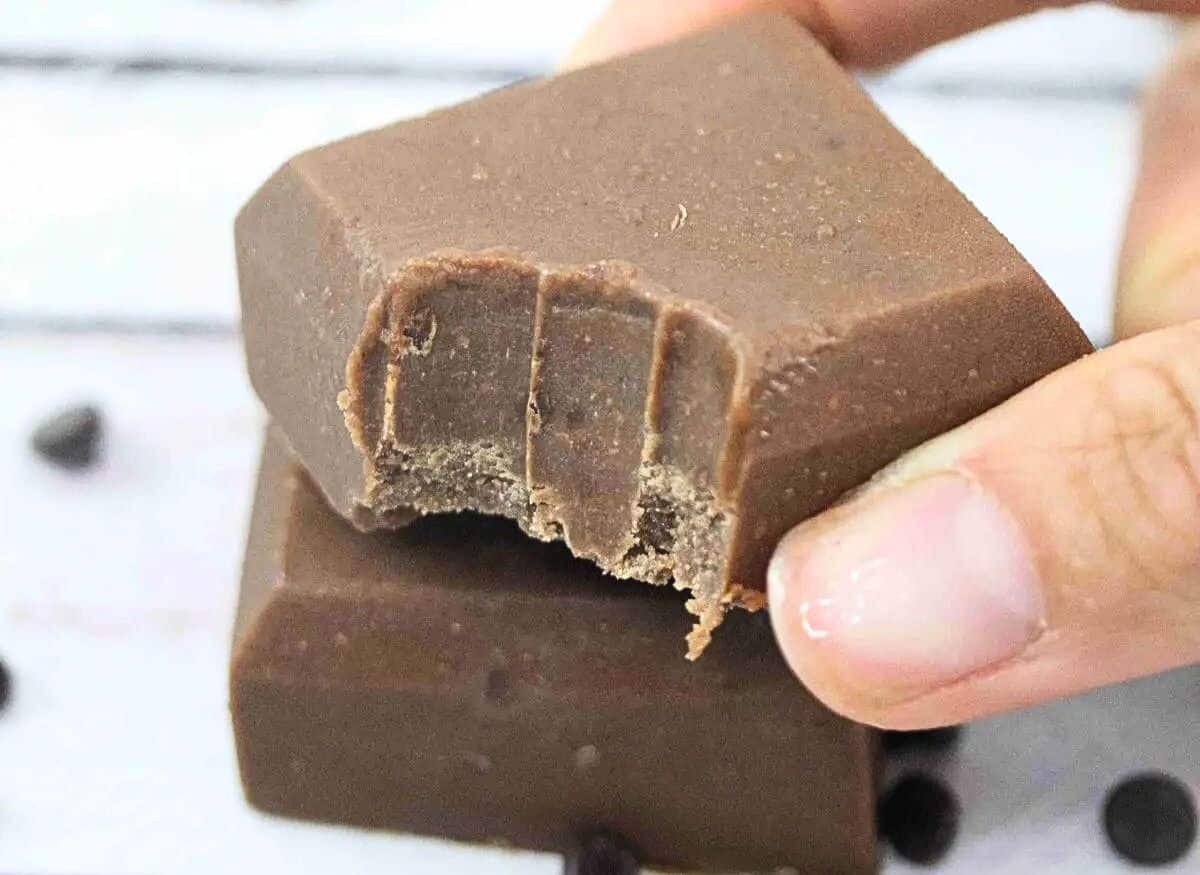 Two squares of chocolate protein fudge on top of each other with the top having a bite out of it and a honey stick behind them.