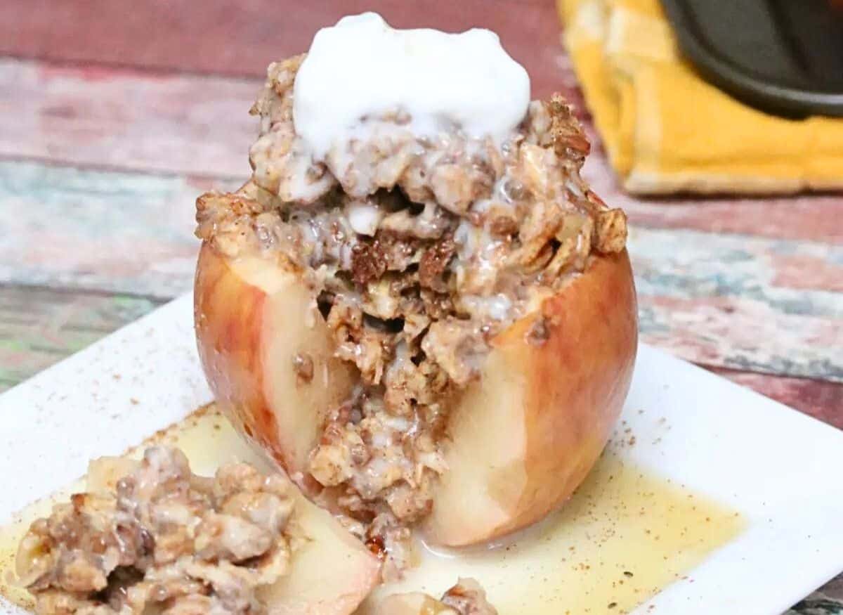 Apple stuffed with oatmeal and topped with whole 30 whipped cream on a white plate with maple syrup on it and more apples in a pan behind them.