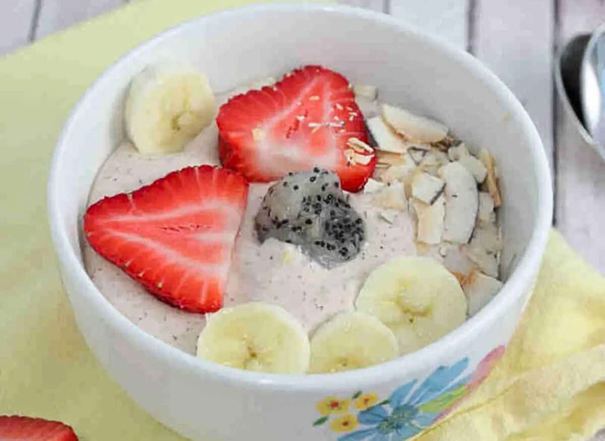 White bowl with a dragon fruit smoothie bowl in it topped with bananas, strawberries, and coconut on a yellow linen with berries, dragon fruit, and spoons around it.