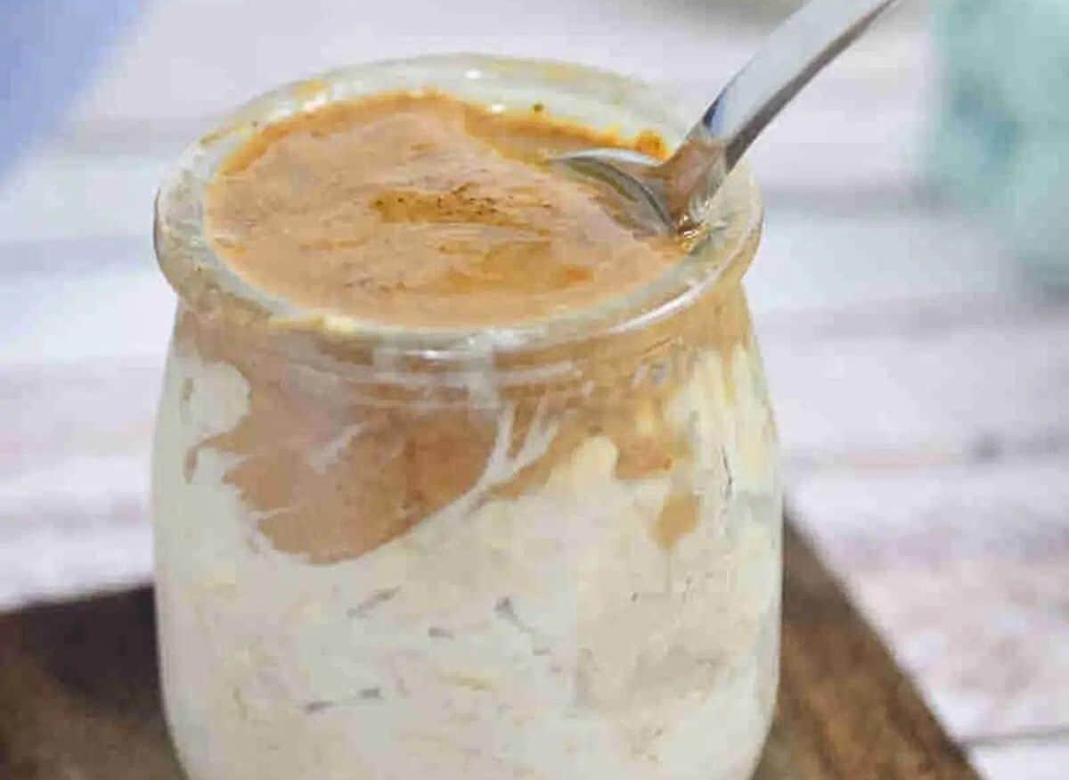 Mason jar with espresso overnight oats topped with almond butter and a spoon in it on a wooden coaster with another jar of oats and a bowl of coffee behind it.