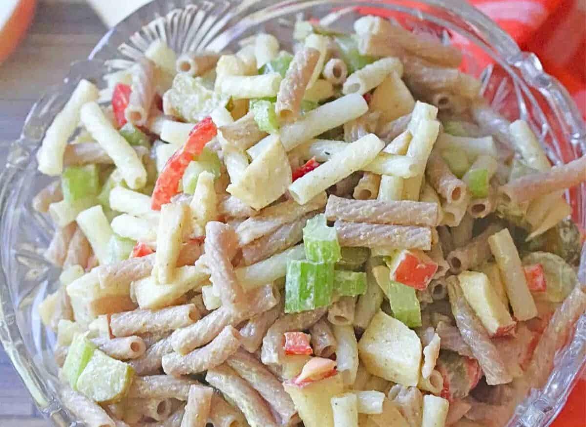 Close up of a glass bowl with macaroni salad in it with celery and red peppers and apple slices and an orange linen behind it.