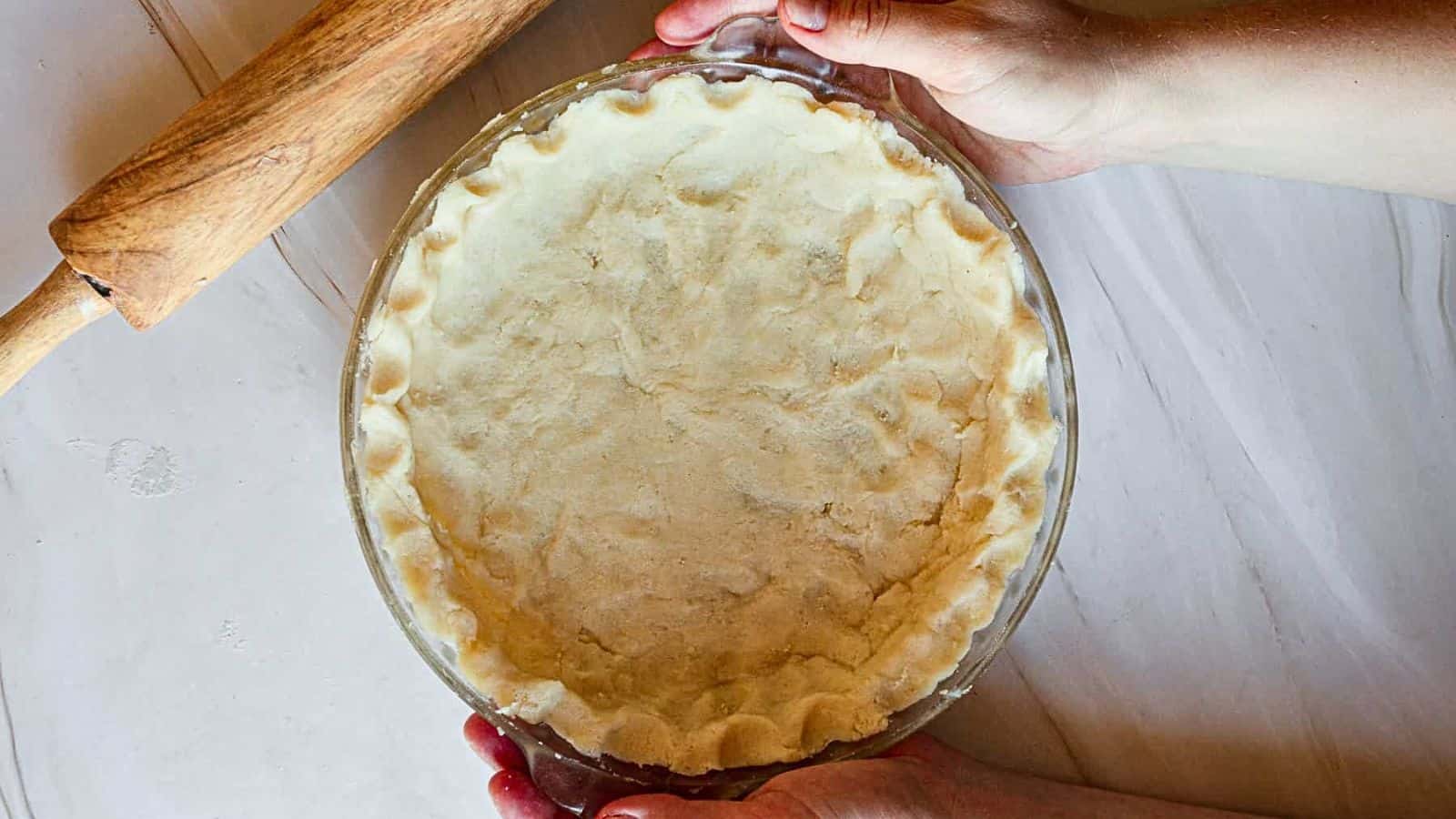A person rolling out a pie crust in a glass pan.