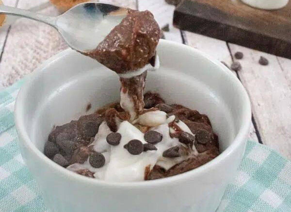 White ramekin on blue checkered linen with chocolate protein pudding and coconut cream in it and a spoon scooping some out.